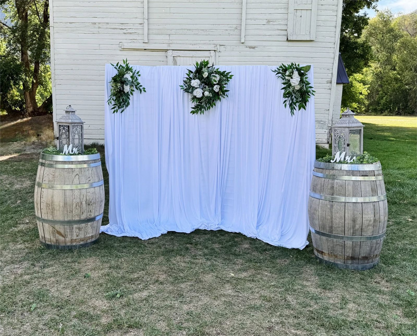 Outdoor wedding ceremony backdrop with white drape, floral swags, whiskey barrels and lanterns in Utah County